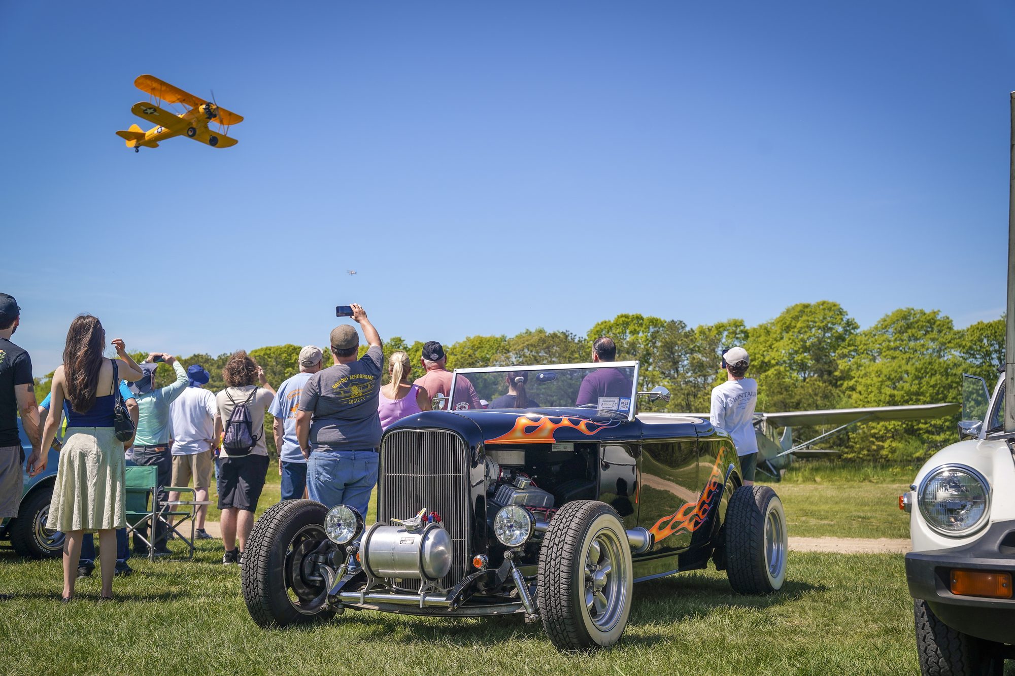 Flamed '32 roadster in the foreground as a crowd watches a yellow biplane fly over